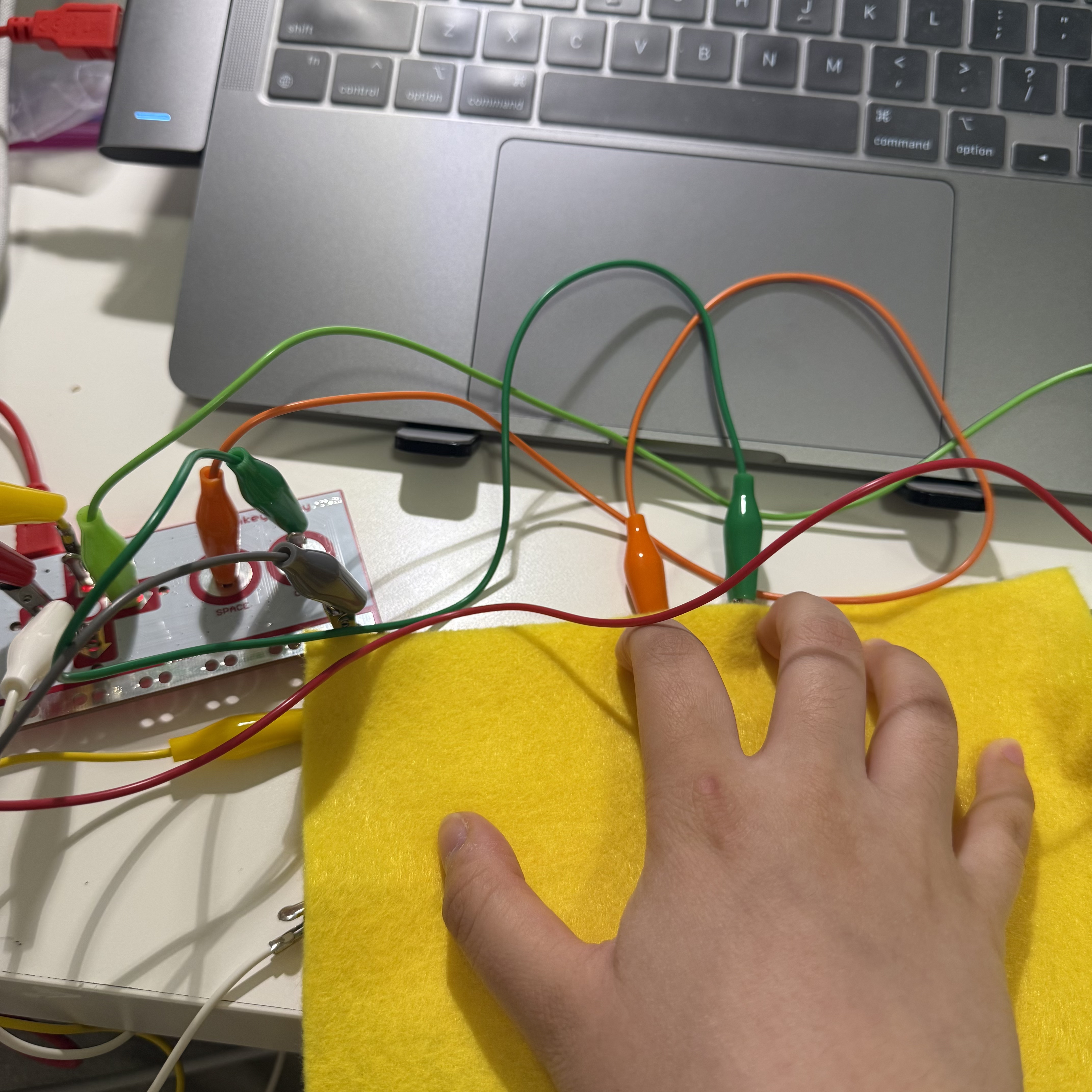 I am demonstrating the functionality of my setup by touching the copper strips on the yellow fabric. This interaction is used to send signals to the Makey Makey board, which then communicates with my laptop. It's a practical way to explore how different materials can be used to control digital applications.
